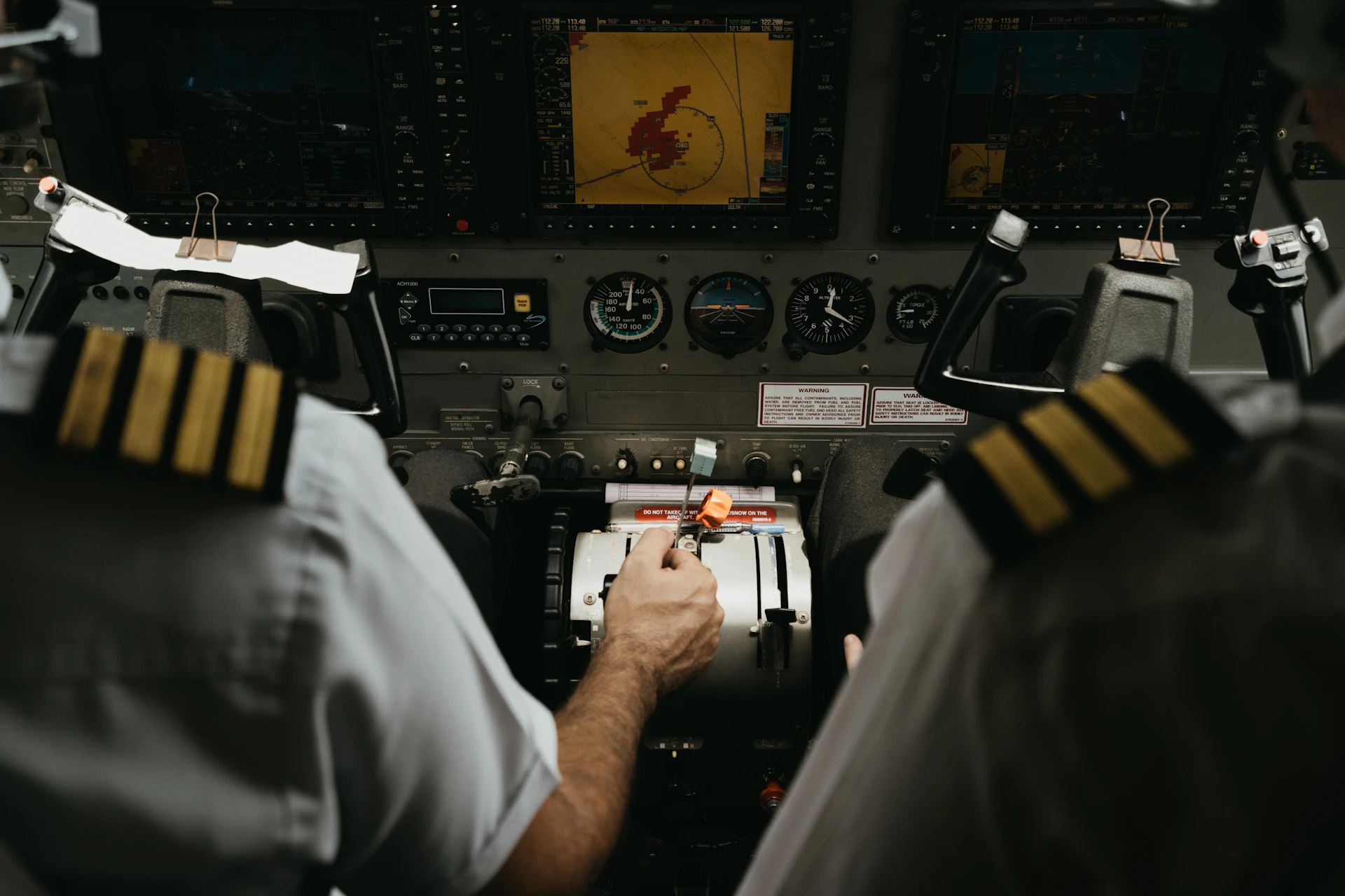 Two pilots sitting in the cockpit of a commercial aircraft, viewed from behind, surrounded by an instrument panel